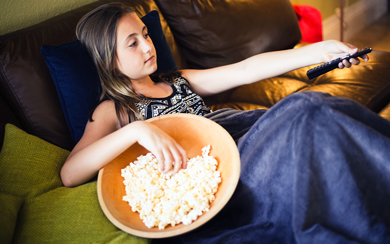 GIrl sits on couch with remote in left hand. Popcorn bowl is placed on couch next to her.