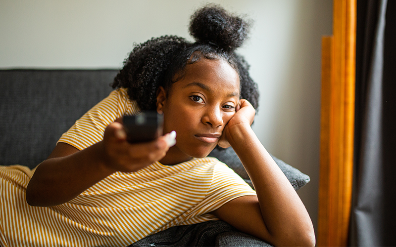 Girl with bored look lay on couch, rests head on left palm, holds TV remote in her right hand