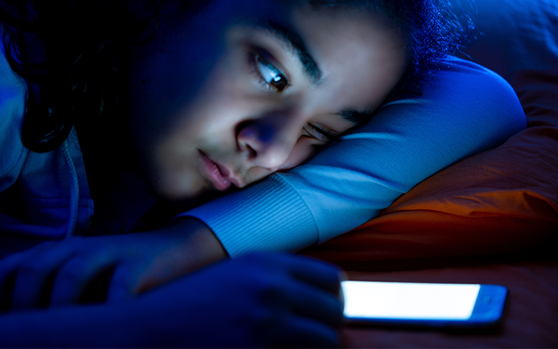 Teen girl lay in bed while staring at her lit-up smartphone on nightstand