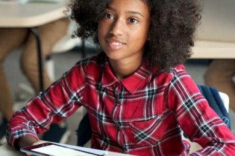 Girl sits at school desk, holding tablet in her hands