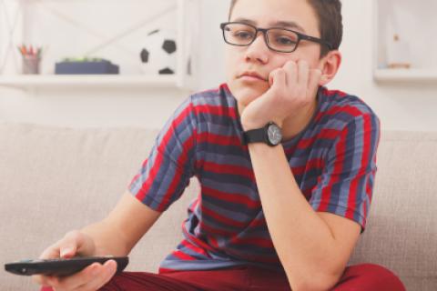 Boy with glasses sits cross-legged on couch, holding TV remote in right hand